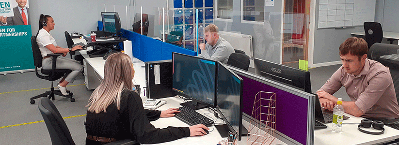 A Tritility office with four employees working at desks with computers, separated by clear partitions.