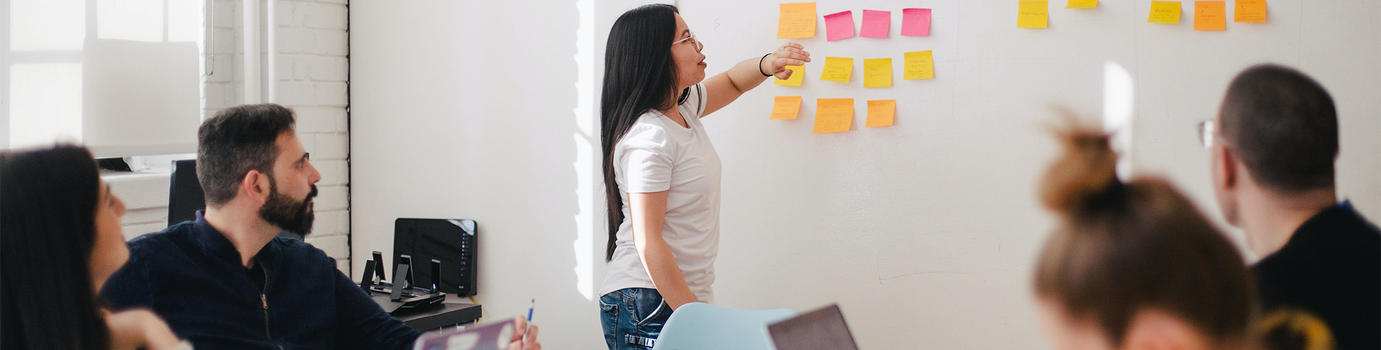 Meeting room with people seated around a table, while a woman stands at the head of the table in front of a whiteboard covered with colourful sticky notes. Attendees are either looking at her or working on their laptops.
