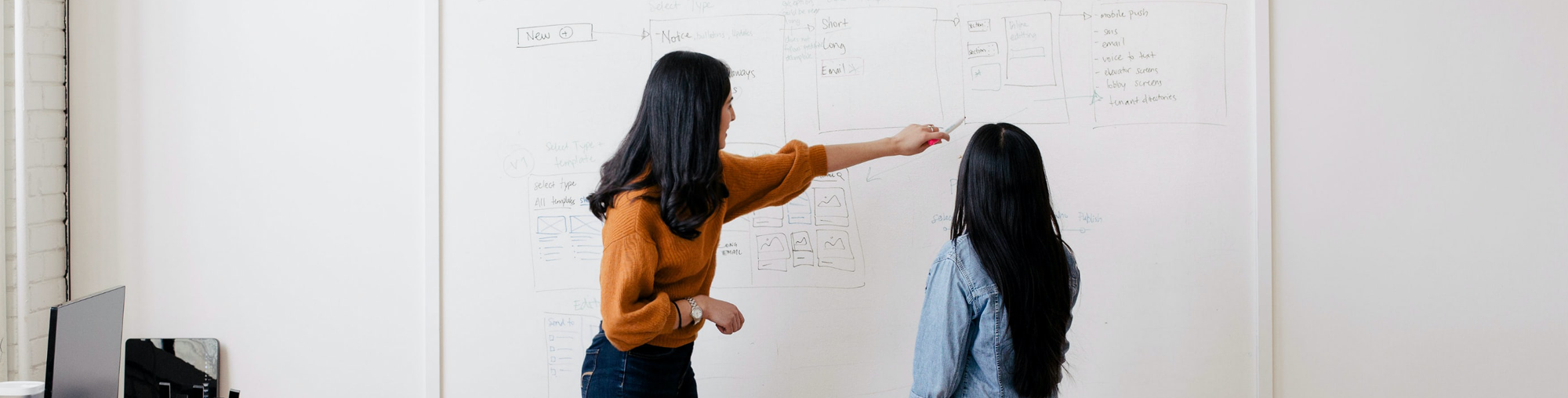 Two women collaborate in front of a whiteboard filled with diagrams and notes, discussing ideas and strategies. A laptop is visible nearby.