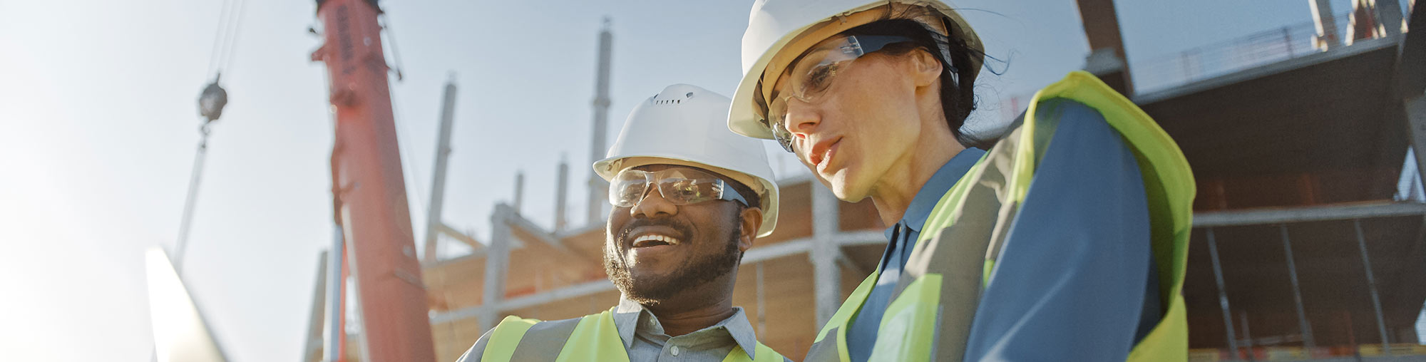 Two construction workers, wearing helmets and safety vests, discuss plans at a building site with cranes and scaffolding in the background.