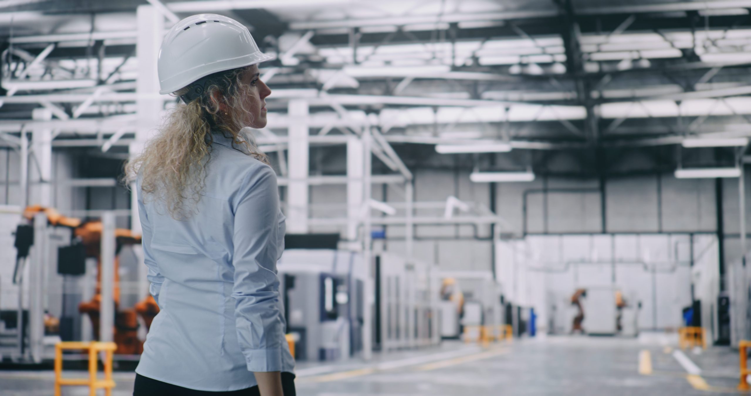 Woman in a blue shirt and white safety helmet considering business energy bills in a large, well-lit interior of a modern industrial facility.