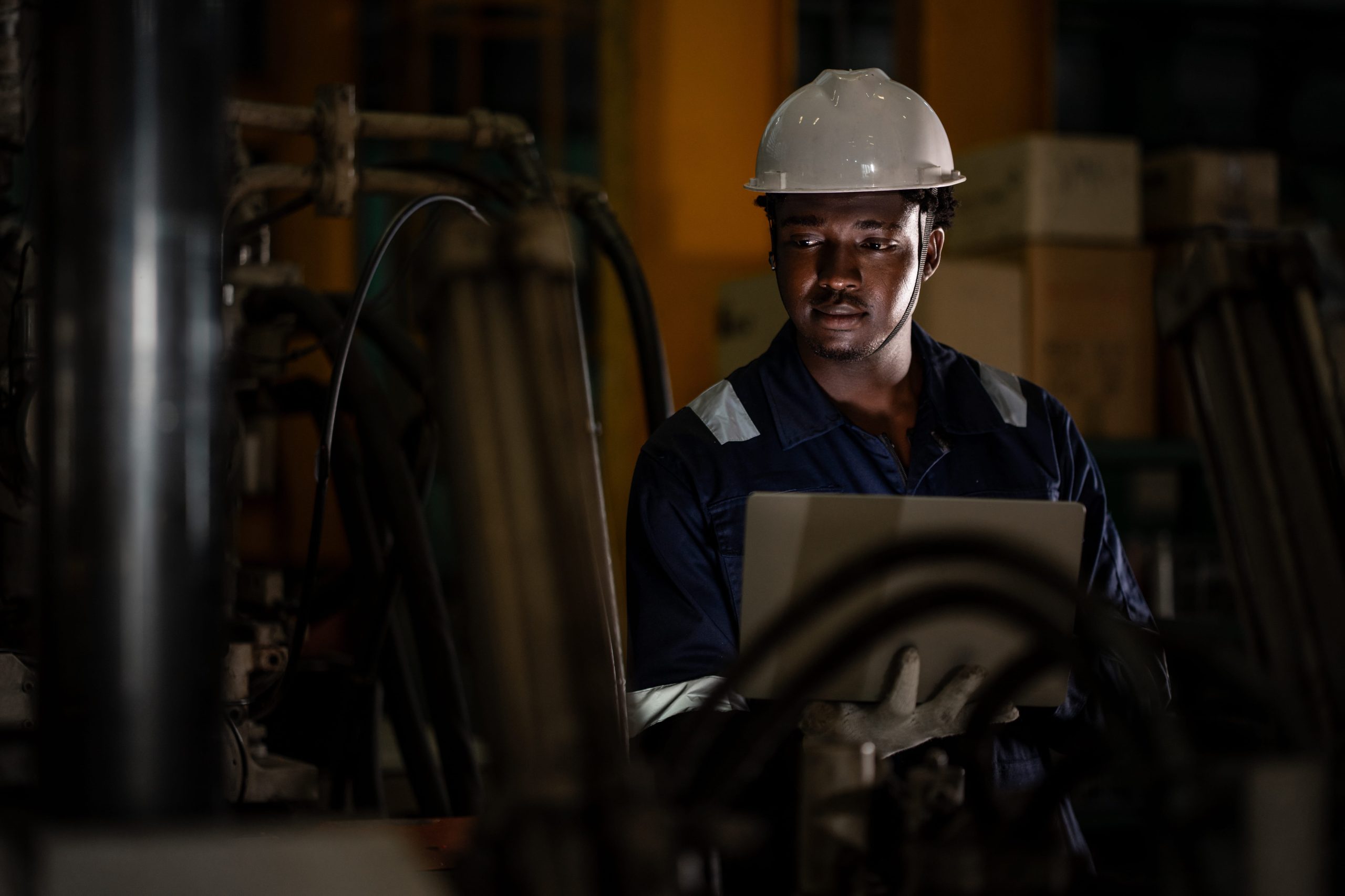 Factory Engineer considers Triad Avoidance in dim lit factory while wearing high vis and looking at laptop